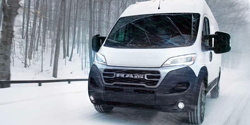 A Ram ProMaster van drives on a snowy road with a forest of bare trees in the background.
