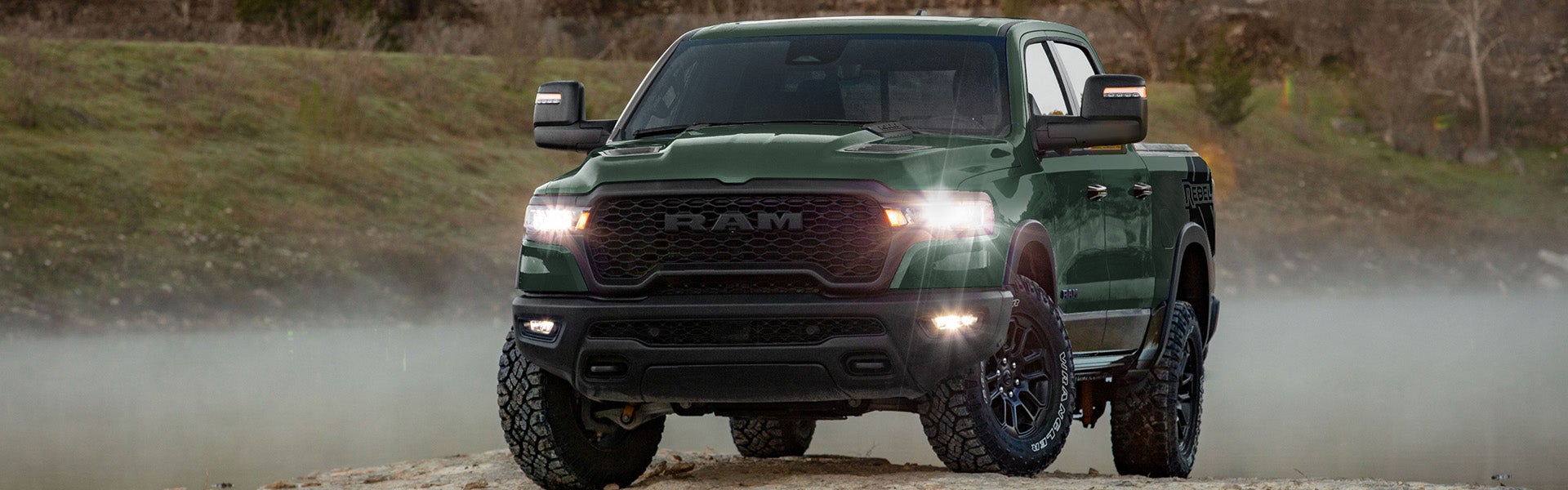 A dark green Ram Rebel pickup truck with glowing headlights and fog lights on a dirt road with dust and fog.