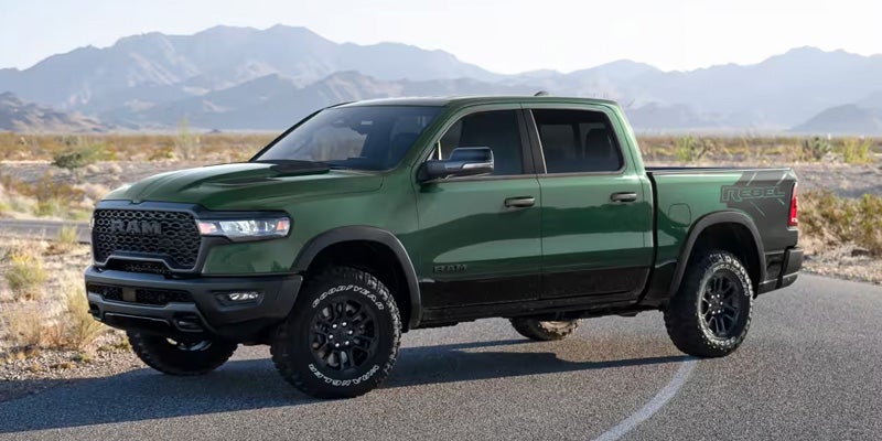 A dark green Ram Rebel pickup truck with black trim and off-road tires, parked on asphalt with mountains in the background.