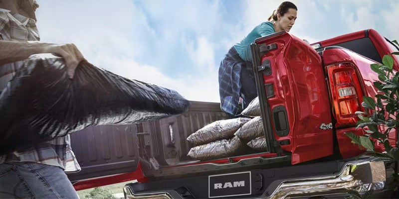 Two people loading bags into the bed of a red Ram pickup truck with green foliage on the right.