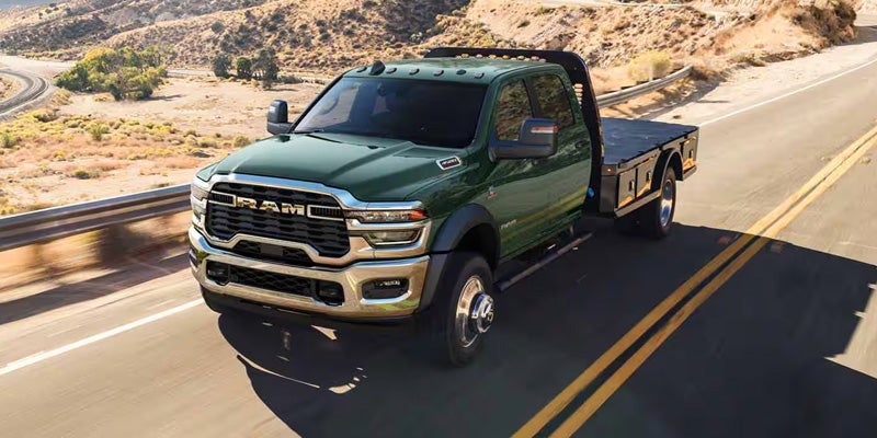 A green Ram flatbed truck drives down a winding desert road with rocky hills and sparse vegetation.
