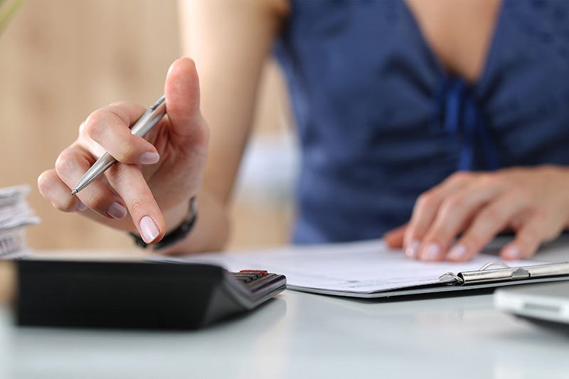 Close-up of a person using a calculator while reviewing documents and writing on a clipboard at a desk