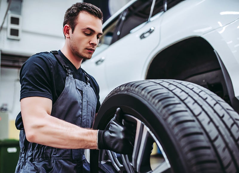 Automotive technician installing or inspecting a vehicle tire and wheel beside a white car in a service workshop