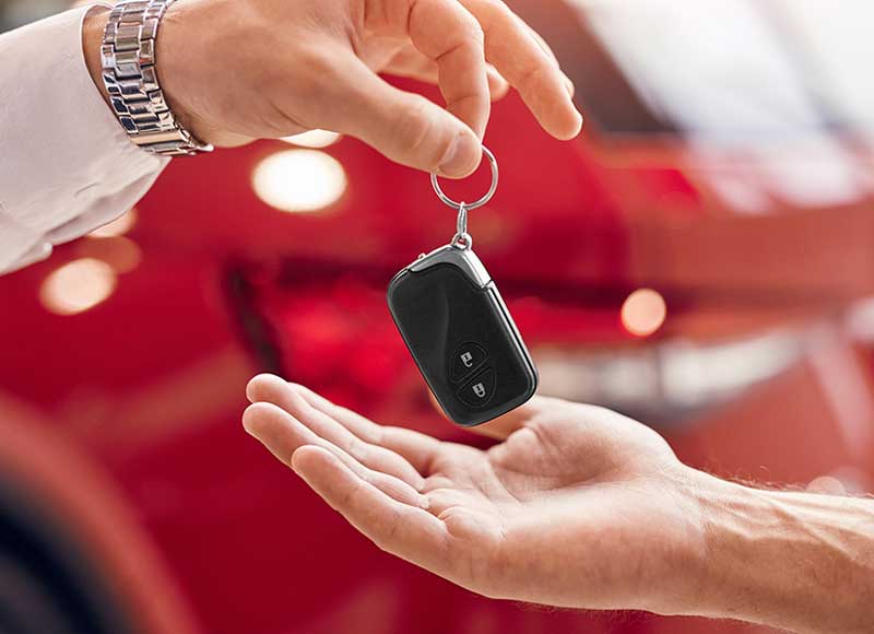 Car key being handed over between two people with a red vehicle blurred in the background, representing vehicle purchase or ownership transfer